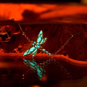 May include: A silver-toned necklace featuring a dragonfly pendant with a glowing turquoise center. The dragonfly design is detailed, with intricate wing patterns. The necklace is displayed on a wooden surface, with a warm, orange-toned background.
