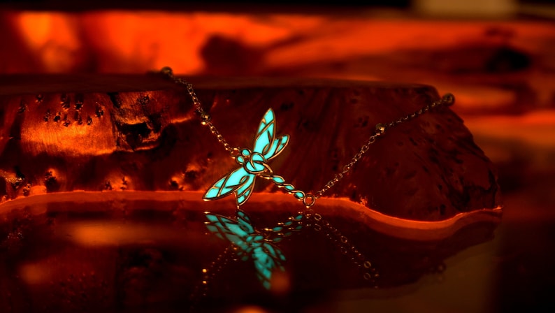 May include: A silver-toned necklace featuring a dragonfly pendant with a glowing turquoise center. The dragonfly design is detailed, with intricate wing patterns. The necklace is displayed on a wooden surface, with a warm, orange-toned background.