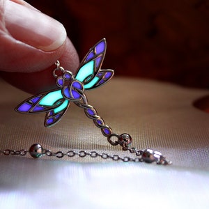 May include: A silver dragonfly pendant necklace with a glowing teal and purple design. The dragonfly's wings are detailed with purple and teal sections, and the body is a twisted silver design. The necklace chain has small silver beads.