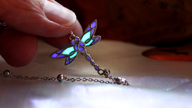 May include: A silver dragonfly pendant necklace with a glowing teal and purple design. The dragonfly's wings are detailed with purple and teal sections, and the body is a twisted silver design. The necklace chain has small silver beads.