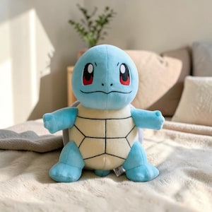 May include: A plush toy of a blue cartoon turtle with large eyes and a beige shell. The toy has a light blue head, arms, and legs. The shell is tan with black lines. The toy is sitting on a soft, light-colored surface.
