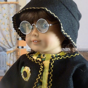 May include: A doll wearing a black felt hat with yellow flower embellishments, round glasses, and a black jacket with yellow trim over a floral print dress.