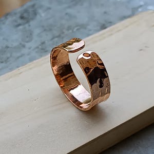 Pode incluir: Um anel ajustável de cobre martelado. O anel tem uma superfície texturizada e um design aberto. A cor cobre é um tom castanho-avermelhado quente. O anel é exibido numa superfície de madeira clara.