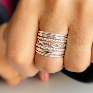 May include: A stack of six silver-toned rings worn on a finger. The set includes two smooth plain bands, two twisted rope-textured bands, and two bands featuring an open infinity or figure-eight link pattern, all arranged in a layered design.