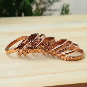 May include: A collection of copper-coloured metal rings featuring various textures and patterns, including braided, floral, and leaf-like designs. The text 925ShreeJaipur is visible on the surface of the rings, which are arranged in a stacked formation.