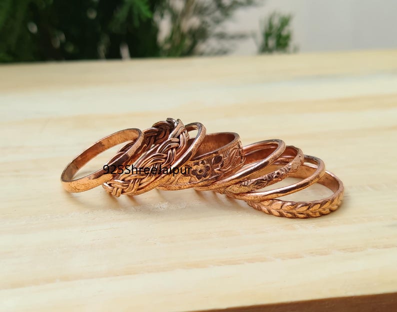 May include: A collection of copper-coloured metal rings featuring various textures and patterns, including braided, floral, and leaf-like designs. The text 925ShreeJaipur is visible on the surface of the rings, which are arranged in a stacked formation.