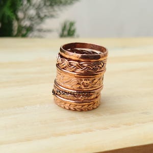 May include: A stack of five copper rings featuring various textures and patterns, including a braided design, a floral motif, and a leaf-like pattern. The text 925ShreeJaipur is visible on the side of the rings. Each ring has a metallic finish.