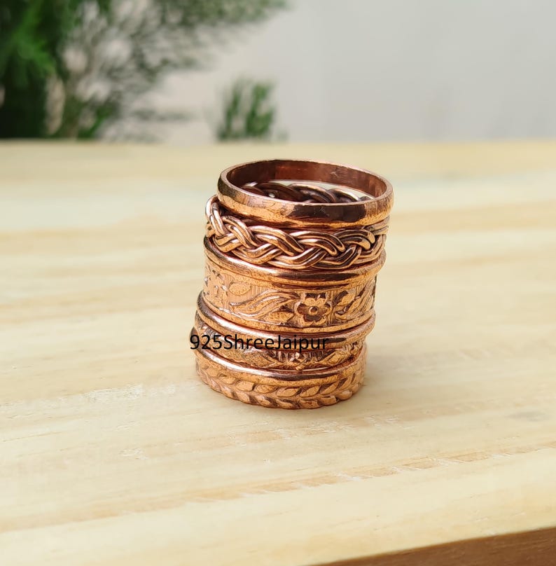 May include: A stack of five copper rings featuring various textures and patterns, including a braided design, a floral motif, and a leaf-like pattern. The text 925ShreeJaipur is visible on the side of the rings. Each ring has a metallic finish.