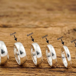 Può includere: Sei anelli color argento lucido disposti in fila, ciascuno etichettato con la propria larghezza: 8mm, 7mm, 6mm, 5mm, 4mm e 3mm. Gli anelli presentano un design a fascia liscia e arrotondata, disposti in ordine decrescente da sinistra a destra.