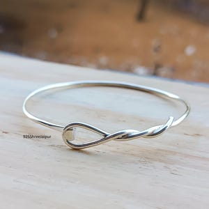 Può includere: Un braccialetto rigido color argento con un design intrecciato. Il braccialetto ha uno stile semplice ed elegante ed è realizzato in metallo lucido. Il braccialetto è su una superficie di legno.