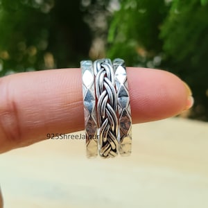 Puede incluir: Un conjunto de tres anillos de plata que presenta una banda central con diseño trenzado flanqueada por dos bandas más delgadas con un patrón de diamantes repetido. El texto 925ShreeJaipur es visible en la superficie de los anillos pulidos.