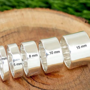 Può includere: Cinque anelli color argento lucido disposti in fila, ognuno con una larghezza diversa. Le larghezze sono etichettate come 3 mm, 5 mm, 8 mm, 10 mm e 15 mm. Gli anelli hanno una superficie liscia e riflettente con un design piatto e largo.