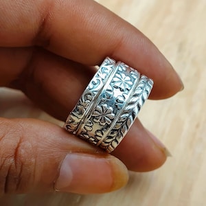Puede incluir: Un anillo de banda ancha de plata con tres secciones giratorias distintas. Las bandas exteriores muestran intrincados patrones de hojas y volutas, mientras que la banda central está decorada con un motivo floral repetitivo en metal pulido.