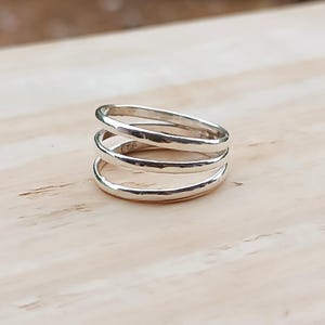 May include: A silver ring with a stacked design. The ring features three bands of varying sizes, creating a layered effect. The bands have a hammered texture, giving the ring a unique, handcrafted appearance. The ring is displayed on a light-colored wooden surface.