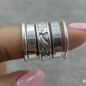 Può includere: Una collezione di anelli d'argento, alcuni con disegni floreali, sono impilati su un dito. Gli anelli variano per larghezza e stile, uno con un motivo floreale e foglia incisa. Il testo "925 Shree Jaipur" è visibile.