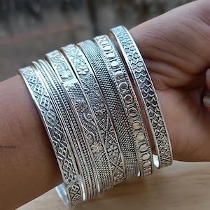 Puede incluir: Una colección de pulseras de plata con intrincados diseños, incluyendo florales, geométricos y texturizados. Las pulseras están apiladas en una muñeca, mostrando la variedad de estilos y la calidad reflectante del metal. La imagen está bien iluminada.