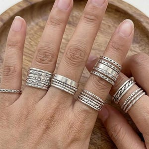 May include: A collection of silver rings in various designs, including bands with beaded, braided, and patterned details. The rings are displayed on multiple fingers, showcasing the different styles and textures. The rings are made of a shiny, metallic material.