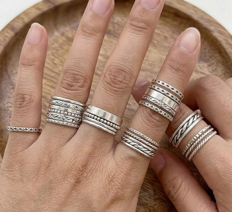 May include: A collection of silver rings in various designs, including bands with beaded, braided, and patterned details. The rings are displayed on multiple fingers, showcasing the different styles and textures. The rings are made of a shiny, metallic material.