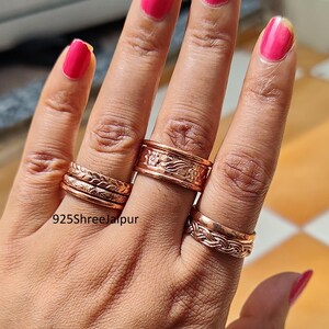 May include: A collection of copper-toned metal rings worn on three fingers. The rings feature various textures, including braided patterns and floral engravings. One ring displays a wider band with a detailed embossed design. The text 925ShreeJaipur is visible.