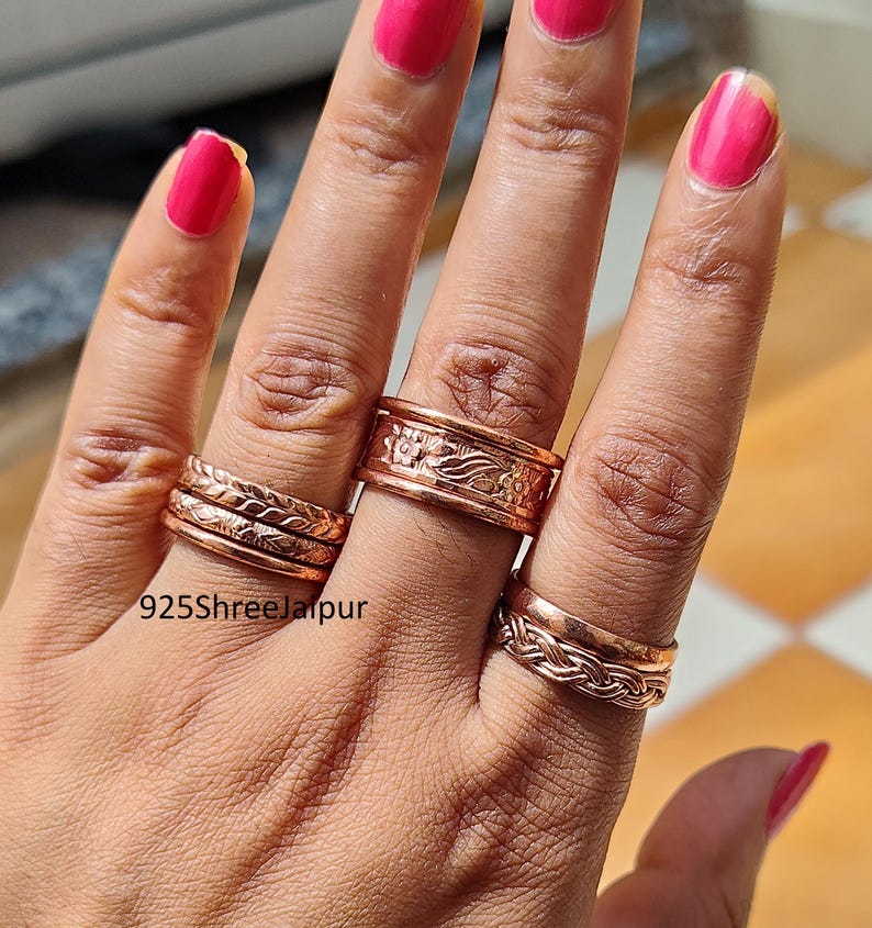 May include: A collection of copper-toned metal rings worn on three fingers. The rings feature various textures, including braided patterns and floral engravings. One ring displays a wider band with a detailed embossed design. The text 925ShreeJaipur is visible.