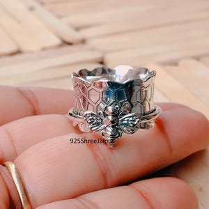 May include: A silver-colored ring with a bee and honeycomb design. The ring features a detailed bee charm and a textured honeycomb pattern around the band. The ring is held by a person.