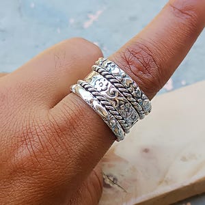 Puede incluir: Un anillo de plata con diseño floral y de cuerda. El anillo tiene múltiples bandas, con patrones detallados de flores y cuerda trenzada. El anillo se lleva en un dedo, mostrando la intrincada artesanía y el diseño de la joyería.