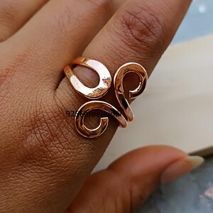 Könnte beinhalten: Ein kupferfarbener Ring mit einem einzigartigen, offenen Design. Der Ring hat drei miteinander verbundene, wirbelnde Formen. Das Metall hat eine polierte Oberfläche, die das Licht reflektiert. Der Ring wird an einem Finger getragen und zeigt seine komplizierten Details. Der Text "925 shreelaxmi" ist sichtbar.