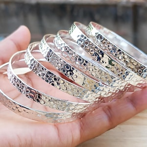 Peut inclure: Plusieurs bracelets argentés avec une texture martelée. Les bracelets sont présentés sur une main, mettant en valeur leur surface réfléchissante. Le design présente une série de petites indentations espacées, créant un effet texturé. Les bracelets sont de couleur argent brillant.