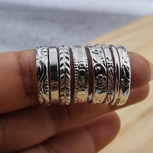 Pode incluir: Um conjunto de sete anéis de prata com várias texturas e padrões, incluindo bandas lisas, motivos de folhas, gravações florais e designs geométricos. Os anéis estão dispostos lado a lado, exibindo um acabamento metálico polido.