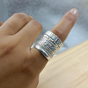 Puede incluir: Un conjunto de cuatro anillos de plata en un dedo. Los anillos presentan texturas distintas, incluyendo una banda lisa, una banda con un patrón grabado fino, una banda con un motivo floral y una banda superior con un patrón geométrico de rombos.