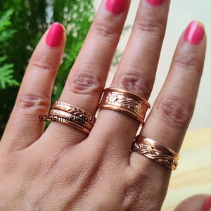 May include: A set of four copper rings worn on three fingers. Two rings on the index finger feature braided and textured patterns. The middle finger ring has a wide band with an embossed floral design. The ring finger band has a braided texture.
