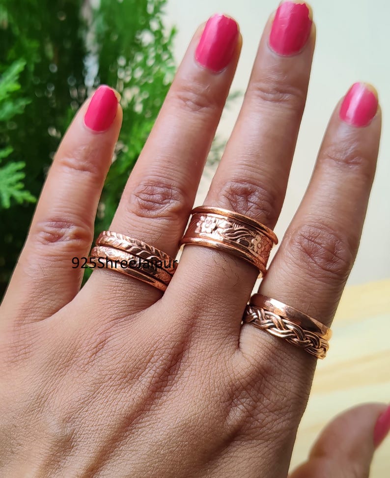 May include: A set of four copper rings worn on three fingers. Two rings on the index finger feature braided and textured patterns. The middle finger ring has a wide band with an embossed floral design. The ring finger band has a braided texture.