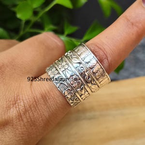 May include: A wide silver spinner ring featuring multiple bands with embossed floral and leaf patterns. The metal surface has an oxidised finish to highlight the intricate botanical details. The text 925ShreeJaipur is visible on the ring.