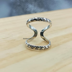 May include: A silver ring featuring two parallel bands connected by a vertical bridge, with a textured diamond pattern along the outer edges of both bands. The text 925ShreeJaipur is printed in black on the inner surface of the ring structure.