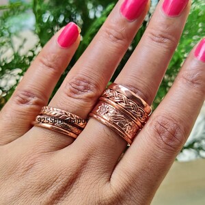 May include: Two copper rings worn on the fingers. The ring on the index finger features a braided band stacked above a wider band with an embossed floral pattern. The ring on the ring finger consists of two thin, textured bands. Text 925ShreeJaipur.