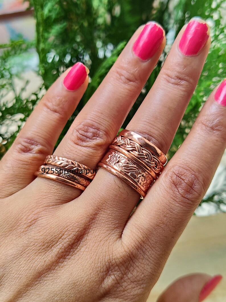 May include: Two copper rings worn on the fingers. The ring on the index finger features a braided band stacked above a wider band with an embossed floral pattern. The ring on the ring finger consists of two thin, textured bands. Text 925ShreeJaipur.