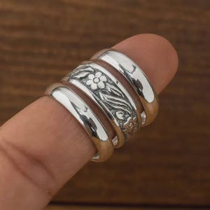 Könnte beinhalten: Ein Satz von vier silbernen Ringen, einer davon mit floralem Design. Die Ringe sind auf einem Finger gestapelt und zeigen ihre polierten, reflektierenden Oberflächen. Der Blumenring hat ein detailliertes Blumen- und Blättermuster.