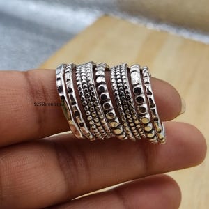 Può includere: Un set di sette anelli in argento con texture varie, inclusi motivi martellati, a perline e punteggiati. Gli anelli sono disposti fianco a fianco, mostrando una finitura metallica. Il testo 925ShreeJaipur è visibile sulla superficie del primo anello.