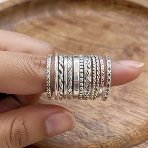 Può includere: Una collezione di anelli d'argento con vari disegni, tra cui intrecciati, floreali e geometrici. Gli anelli sono impilati su un dito, mostrando le diverse trame e stili. Gli anelli sono realizzati in un materiale metallico lucido.