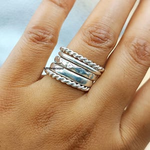 Peut inclure: Une pile de quatre bagues en argent comprenant deux anneaux torsadés, un anneau lisse poli et un anneau avec une texture à motif losange. Le texte 925ShreeJaipur est visible sur la surface de l'anneau lisse au centre de la pile.