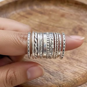 May include: A collection of silver rings with various designs, including braided, floral, and geometric patterns. The rings are stacked on a finger, showcasing the different textures and styles. The rings are made of a shiny, metallic material.