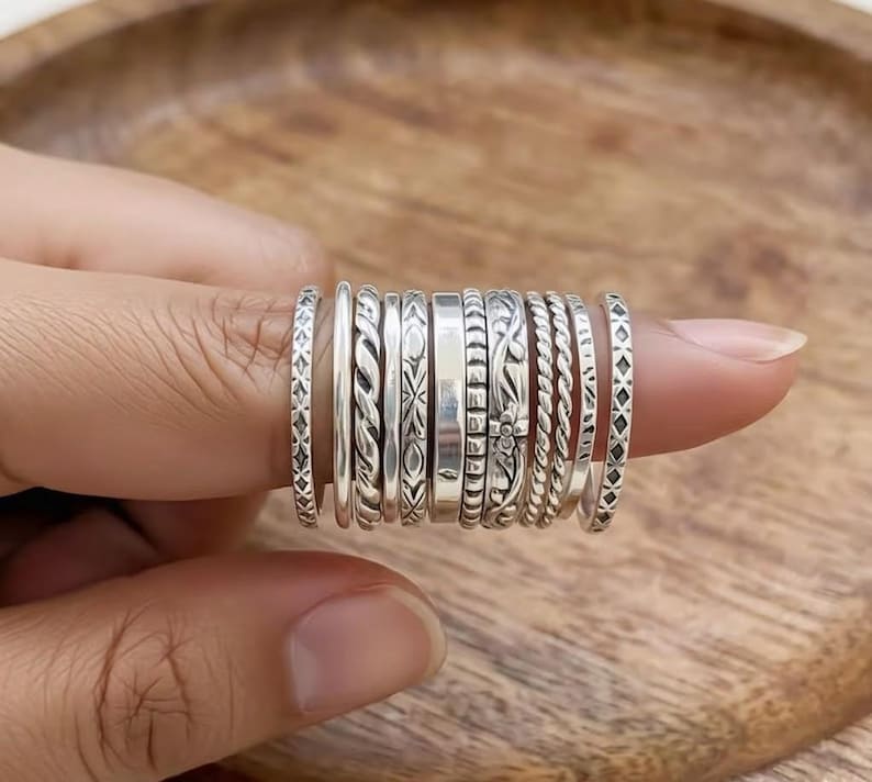 May include: A collection of silver rings with various designs, including braided, floral, and geometric patterns. The rings are stacked on a finger, showcasing the different textures and styles. The rings are made of a shiny, metallic material.
