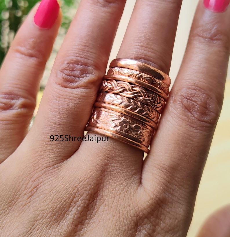 May include: A stack of five copper rings featuring various textures, including a smooth band, a geometric pattern, a braided design, a twisted rope style, and a wide band with an engraved floral motif. The text 925ShreeJaipur is visible.