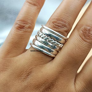 May include: A stack of five silver rings featuring varied textures, including smooth bands, a twisted rope design, and a faceted band. The text 925ShreeJaipur is visible on the surface of the bottom ring. The set is worn together on a finger.