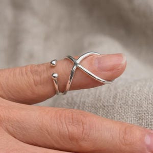Könnte beinhalten: Ein silberfarbener Ring mit einem X-förmigen Design und zwei kleinen Kugelakzenten. Der Ring wird an einem Finger getragen und unterstreicht seinen modernen und minimalistischen Stil. Das Design des Rings ist ein einzigartiges und auffälliges Schmuckstück.