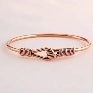 Puede incluir: Una pulsera de brazalete de oro rosa con un diseño de nudo único. La pulsera presenta una banda lisa y circular con un cierre en forma de nudo. El cierre está envuelto con detalles de alambre, agregando textura e interés visual a la joyería.
