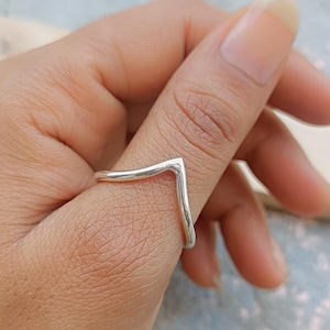 May include: A silver-colored ring with a V-shaped design. The ring is worn on a finger, showcasing its minimalist and modern aesthetic. The ring's smooth, polished surface reflects light, highlighting its clean lines and simple elegance.