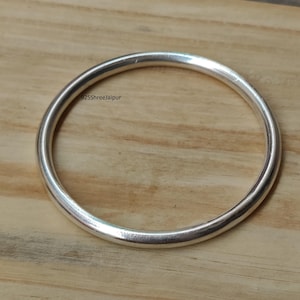 Puede incluir: Una pulsera de plata lisa y pulida con forma circular. La superficie metálica es reflectante y de grosor uniforme. El texto 925ShreeJaipur está impreso en pequeñas letras negras sobre la superficie de la pulsera de plata.