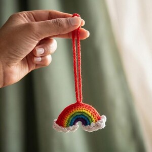 May include: A crocheted rainbow decoration featuring red, orange, yellow, green, blue, and purple arches. The rainbow has white fluffy clouds at the base and a red hanging string. The ornament is held up by a hand.