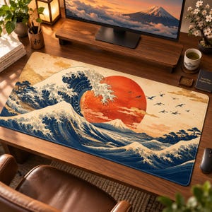 Puede incluir: Una alfombrilla de escritorio rectangular con una ilustración estilizada de una gran ola azul, un sol rojo prominente y un pico de montaña. El diseño incluye espuma de mar blanca, agua azul oscuro y aves sobre un fondo de color crema.